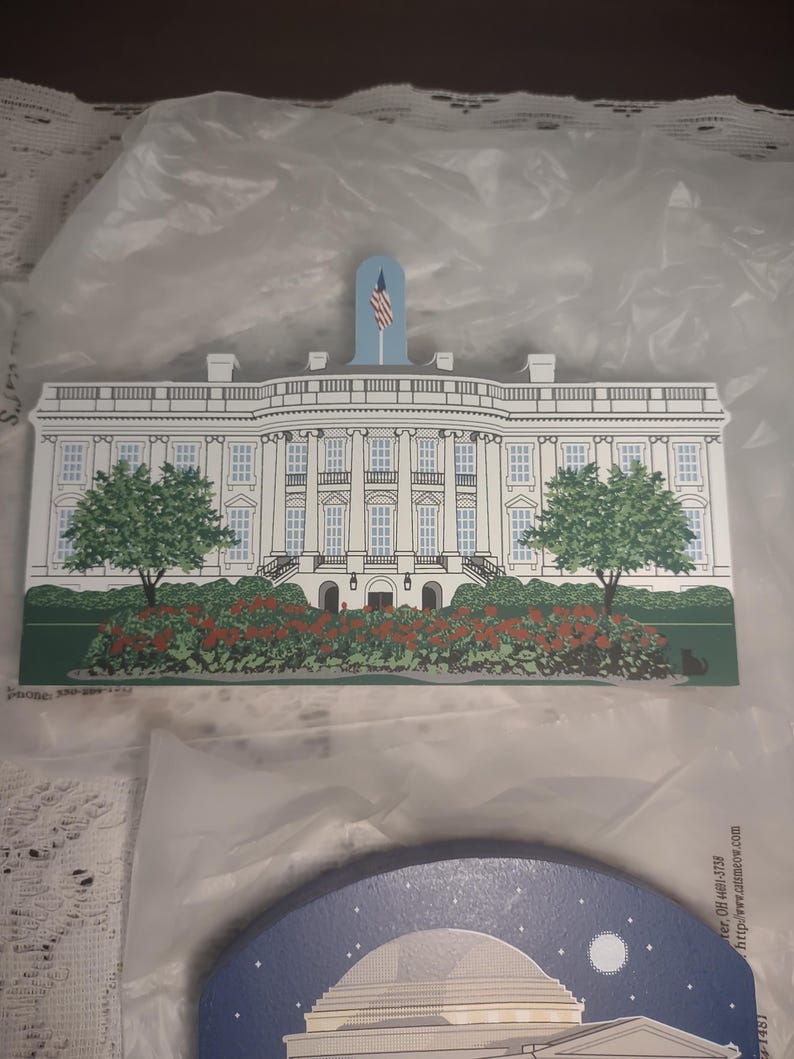 May include: A wooden White House cutout with a blue flag and an American flag. The building is white with black windows, green trees, and red flowers. The cutout is on a white surface.
