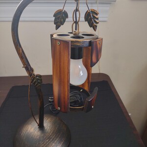 May include: A decorative table lamp with a bronze finish and a wooden shade. The lamp features a curved arm and a base with a circular design. The shade is made of wooden panels and has a light bulb inside. The lamp is designed with leaf accents.