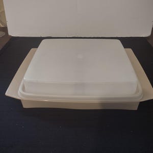 May include: A square, off-white butter dish with a clear, square lid. The dish sits on a rectangular base, also off-white. The lid has a small, circular handle. The background is a dark surface and a white wall.