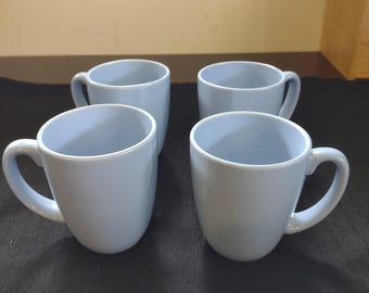 May include: Four light blue ceramic mugs with curved handles are arranged on a dark surface. The mugs have a simple, rounded design and a smooth, matte finish. The mugs are empty and ready for use.
