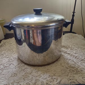 May include: A large, stainless steel cooking pot with a lid and black handles. The pot is cylindrical with a shiny, reflective surface. The lid has a black knob. The pot is sitting on a white lace tablecloth.