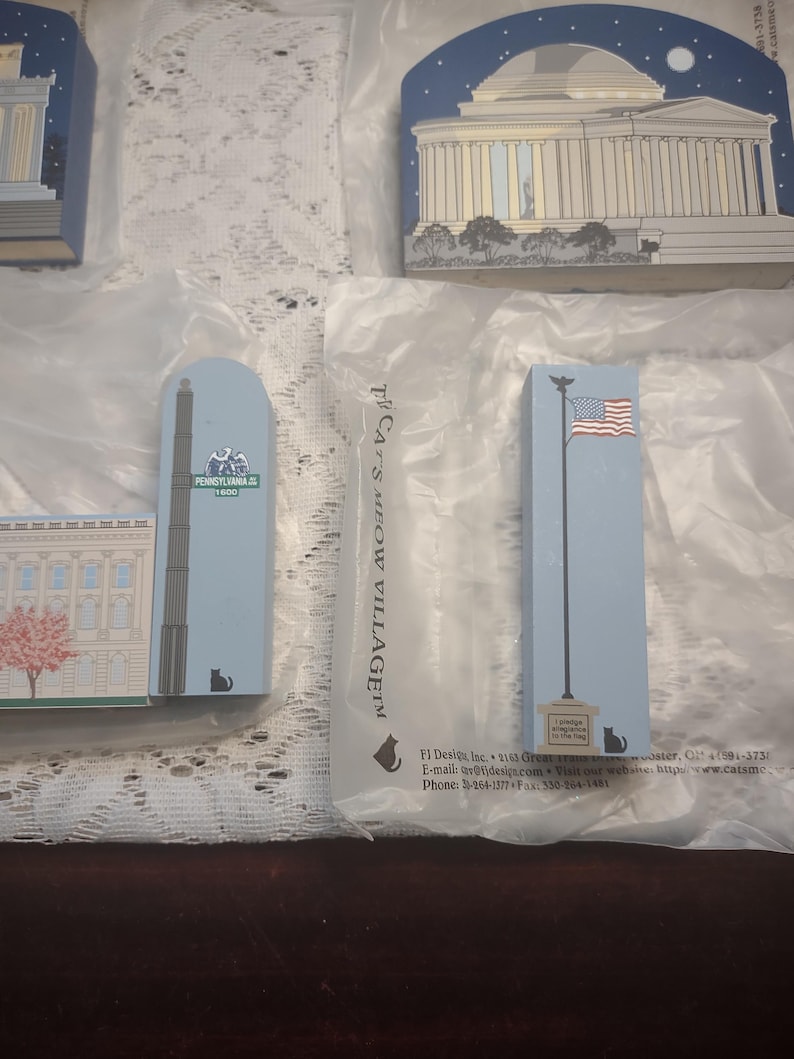 May include: A collection of rectangular wooden ornaments featuring iconic American landmarks. The ornaments are painted in various colors and designs, including the U.S. Capitol building, a tall obelisk, and a flagpole with the American flag. The text "PENNSYLVANIA 1600" is visible.