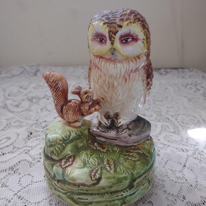 May include: A ceramic owl figurine with a squirrel perched on a green base. The owl has brown and white feathers, with red accents around the eyes. The squirrel is brown. The base is a detailed, green, leaf-covered design.