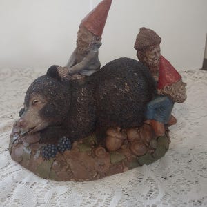 May include: A whimsical figurine featuring a brown bear with two gnomes. One gnome wears a red hat and sits atop the bear, while the other gnome, also in a red hat, sits on the side. The base includes acorns and berries.