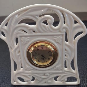 May include: A white ceramic mantel clock with a gold-tone clock face. The clock has Roman numerals and a decorative, openwork design featuring floral and scroll motifs. The clock is approximately 6 inches tall.