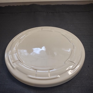 May include: A round, off-white lid with a raised, patterned edge. The lid has a smooth, glossy finish and appears to be made of ceramic or a similar material. The design includes rectangular and curved elements.
