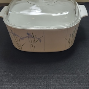 May include: A square white ceramic casserole dish with integrated side handles and a clear glass lid. The exterior features a light beige base color decorated with a pattern of purple iris flowers and green stems arranged in vertical lines.