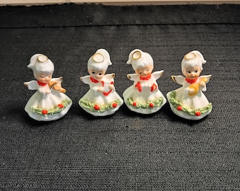 May include: Four ceramic angel figurines, each wearing a white dress with a green garland and red berries at the hem. Each angel has a gold halo, red collar, and white wings, and holds a different musical instrument: a violin, a harp, a horn, and a lute.