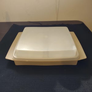 May include: A beige square plastic container with a clear, square lid. The container has a slightly raised edge and is sitting on a dark blue surface. The lid has a small circular indentation in the center.