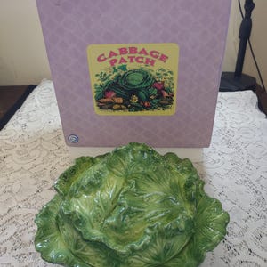 May include: A green ceramic cabbage-shaped dish with a lid, sitting in front of a box with a "Cabbage Patch" logo. The dish is detailed with leaf textures and a glossy finish. The box is light purple with a diamond pattern.