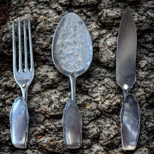 May include: A set of antique cutlery comprising a fork, spoon, and knife. The silver-toned metal utensils are displayed on a textured, dark brown surface. The spoon's bowl has a hammered design.