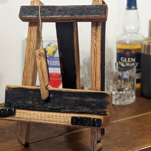 May include: A small wooden easel crafted from reclaimed whiskey barrel staves. The easel features a dark brown stain and black accents. It is resting on a wooden surface.