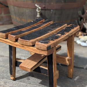 May include: A small wooden stool crafted from reclaimed barrel staves. The seat is constructed from several black-stained staves, and the legs are made from natural wood.