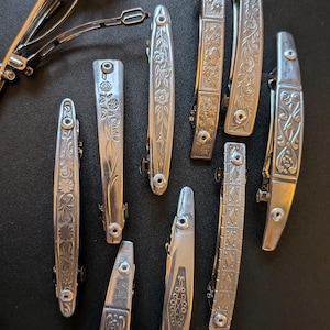 May include: A collection of nine silver-toned metal hair barrettes featuring various engraved patterns including floral, geometric, and scrollwork designs. Each barrette has a spring-loaded clasp mechanism visible on the back of the open piece.