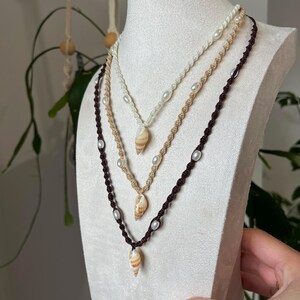 May include: Three macrame necklaces with seashell pendants and pearl accents displayed on a white mannequin. The necklaces are in shades of white, beige, and brown. The seashells are tan and brown with a spiral pattern.