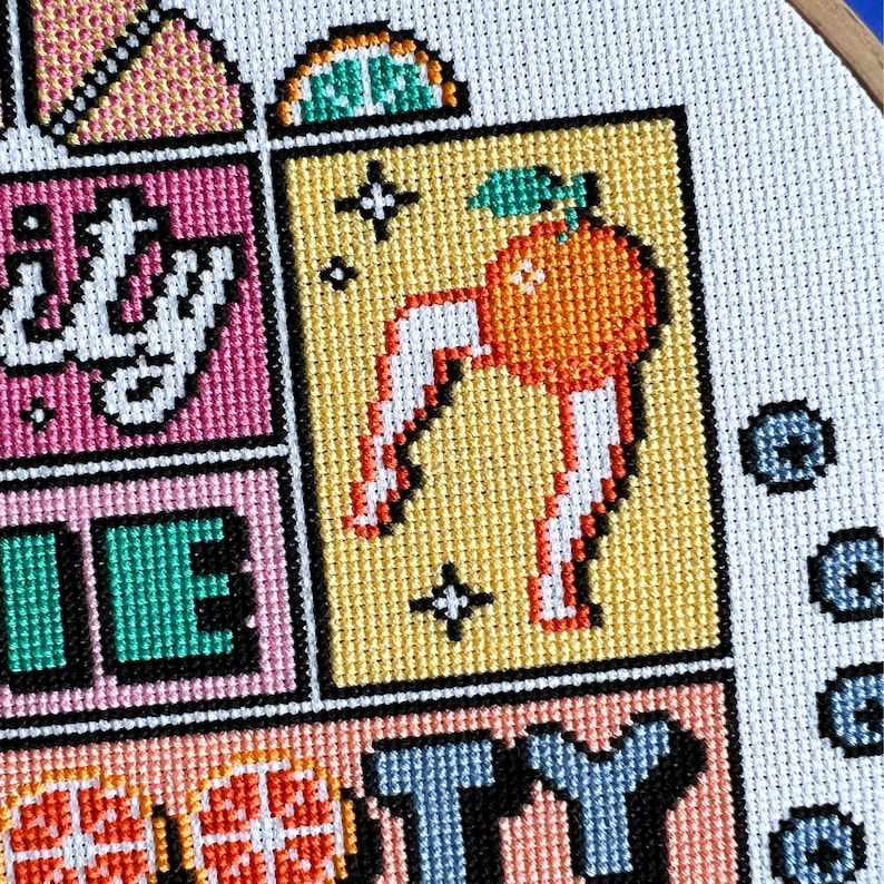 May include: Close-up of a cross-stitch design with a vibrant colour palette. The design features a pixelated orange with white legs and a green leaf, set against a yellow background. Other blocks display text and geometric shapes.
