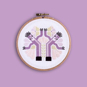 Può includere: Un telaio da ricamo a punto croce con un disegno simmetrico di due figure viola con capelli e piedi neri e braccia tese. Le figure sono ambientate su uno sfondo di elementi floreali rosa e gialli, il tutto all'interno di un telaio di legno.