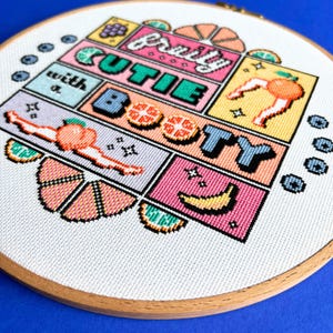 May include: A cross-stitch design in a wooden hoop, displaying the text "fruity cutie with a booty". The colourful design incorporates fruit illustrations, including a peach, banana, and orange segments, in a comic book style.
