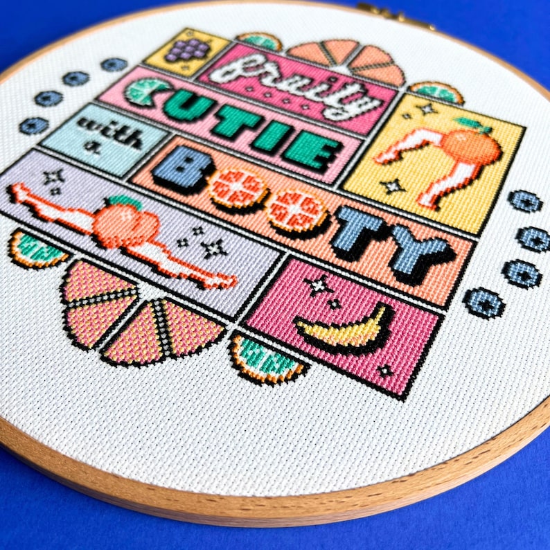 May include: A cross-stitch design in a wooden hoop, displaying the text "fruity cutie with a booty". The colourful design incorporates fruit illustrations, including a peach, banana, and orange segments, in a comic book style.