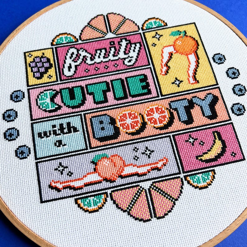 May include: A cross-stitch hoop with the words "Fruity Cutie Booty" and fruit designs. The design includes a grape cluster, an orange with legs, watermelon slices, a peach, and a banana. The text and images are in various colours.