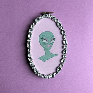 Peut inclure: Broderie au point de croix d'un visage d'extraterrestre vert avec de longs cils et des lèvres roses, sur fond rose clair. Le cadre ovale est orné de cristaux transparents et étincelants. L'œuvre est montée dans un cercle à broder en bois.
