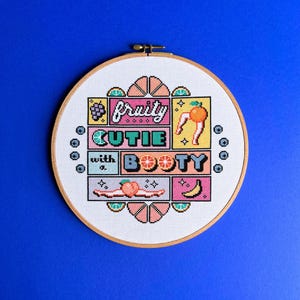 May include: A cross-stitch embroidery hoop featuring the phrase "fruity cutie with a booty" in a colourful, pixelated design. The design incorporates fruit illustrations like grapes, oranges, peaches, and bananas on white fabric, set against a blue background.