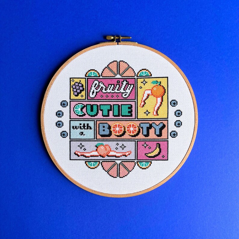 May include: A cross-stitch embroidery hoop featuring the phrase "fruity cutie with a booty" in a colourful, pixelated design. The design incorporates fruit illustrations like grapes, oranges, peaches, and bananas on white fabric, set against a blue background.