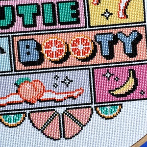 May include: A cross-stitch design with the words "Cutie Booty" in block letters. The design includes images of a grapefruit, peach, and banana. The colour scheme features pink, green, yellow, and blue. The design is on a white background.