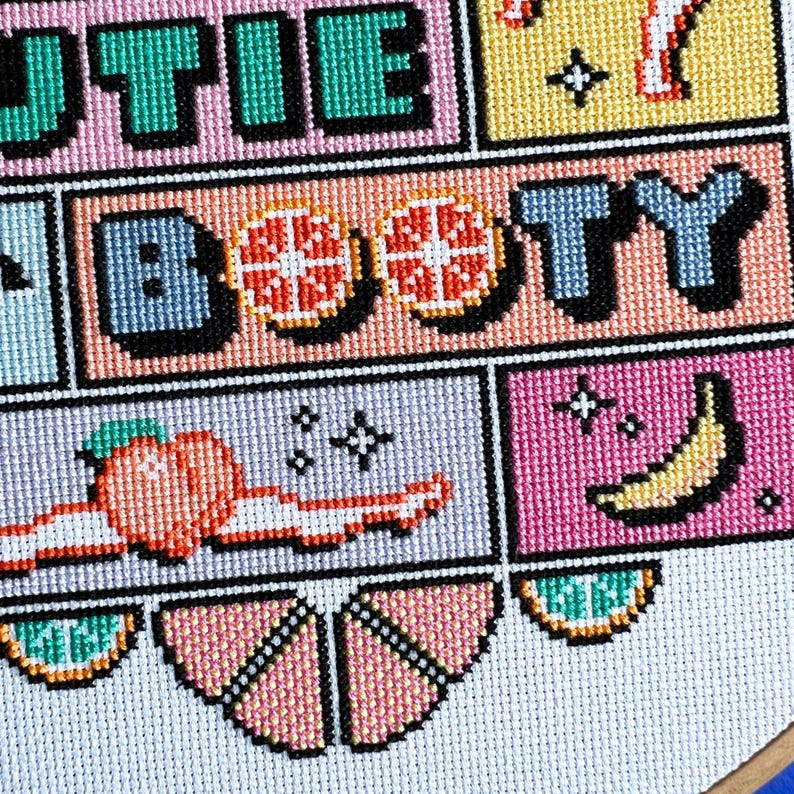 May include: A cross-stitch design with the words "Cutie Booty" in block letters. The design includes images of a grapefruit, peach, and banana. The colour scheme features pink, green, yellow, and blue. The design is on a white background.