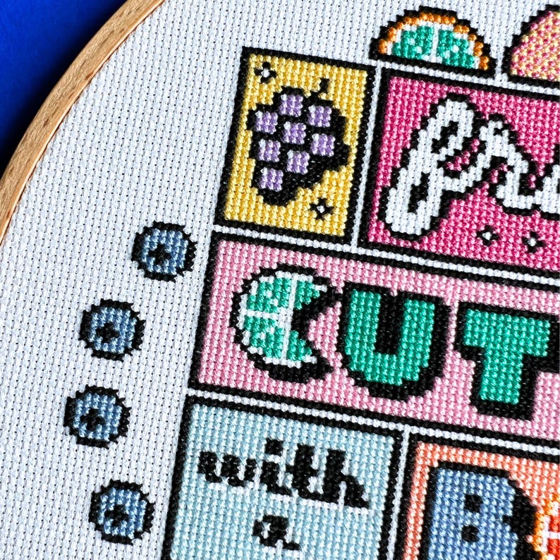 May include: Cross-stitch embroidery in a wooden hoop. The design features colourful blocks with text and images, including a bunch of grapes, a slice of watermelon, and the word "CUT". The colour palette includes pink, yellow, green, and blue.