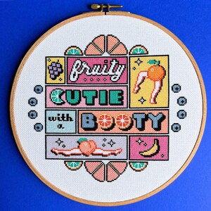 May include: A cross-stitch embroidery in a wooden hoop features the text "fruity cutie with a booty" with fruit illustrations. The design includes a grape bunch, an orange with legs, a peach, and a banana. The background is white with a blue border.