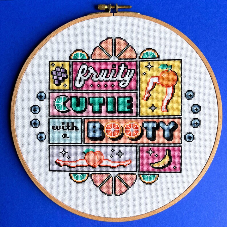 May include: A cross-stitch embroidery in a wooden hoop features the text "fruity cutie with a booty" with fruit illustrations. The design includes a grape bunch, an orange with legs, a peach, and a banana. The background is white with a blue border.
