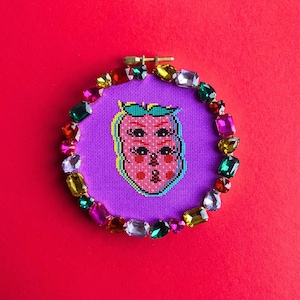 Puede incluir: Un bastidor de bordado de punto de cruz con una cara de fresa rosa con múltiples ojos y un contorno amarillo, sobre un fondo morado. El bastidor está adornado con gemas de colores, sobre un fondo rojo.