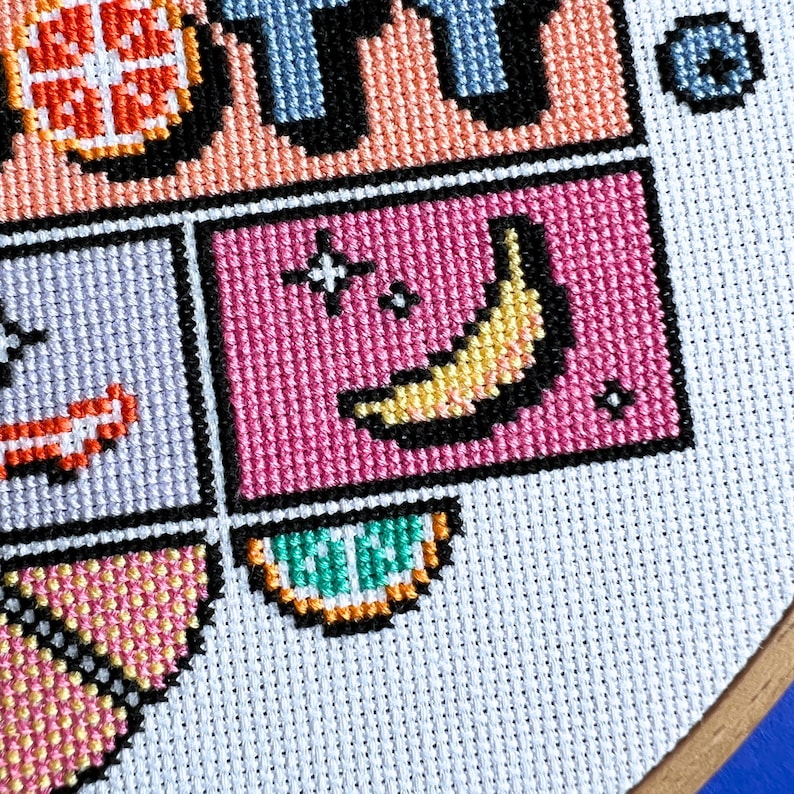 May include: Close-up of a cross-stitch design featuring a banana on a pink background. The design is composed of various colourful squares, including a section with a yellow banana, black outlines, and white cross-stitch fabric.