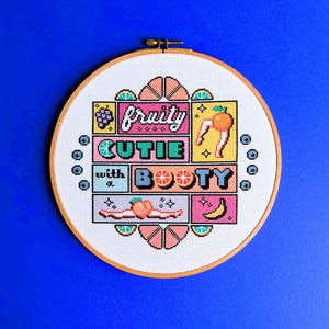 May include: A cross-stitch embroidery hoop featuring the phrase "fruity cutie with a booty" in a colourful, pixelated design. The design incorporates fruit illustrations, including grapes, a peach, and a banana, on a white fabric background.