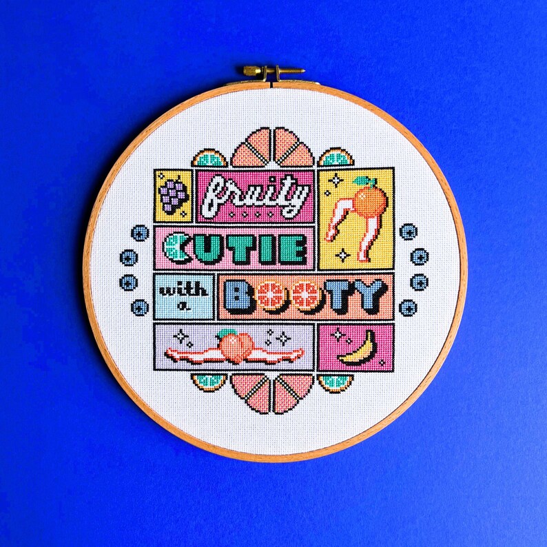 May include: A cross-stitch embroidery hoop featuring the phrase "fruity cutie with a booty" in a colourful, pixelated design. The design incorporates fruit illustrations, including grapes, a peach, and a banana, on a white fabric background.
