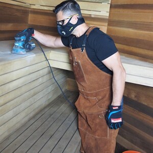 May include: A person wearing a black shirt, safety glasses, and a respirator is using a teal and black Makita sander on wooden planks. The person is wearing a brown leather apron and black and blue work gloves.