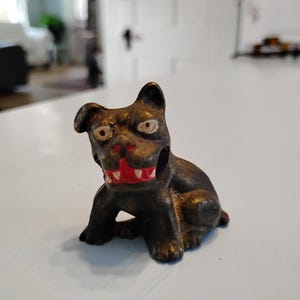 Peut inclure: Une petite figurine de chien en fonte noire avec des yeux et une bouche peints en rouge. Le chien est assis, les pattes repliées sous le corps.