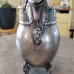May include: Antique silver-colored tea pot with ornate details. The pot features a rounded body, a spout with a face design, and decorative relief around the top. It stands on three ornate feet and has a lid with a handle.