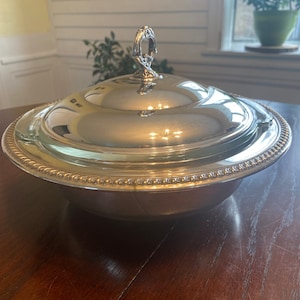 Puede incluir: Un bol de servir plateado con tapa y un asa decorativa. El bol tiene un borde con cuentas y una superficie lisa y reflectante.