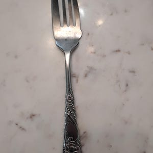 Könnte beinhalten: Eine silberfarbene Metallgabel mit einem vierzinkigen Kopf, bei dem die linke Zinke abgebrochen ist. Der Griff ist mit einem detaillierten, geprägten Blumen- und Rankenmuster verziert, das sich vom Hals bis zum abgerundeten Ende erstreckt.
