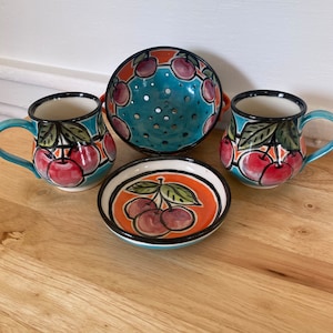 May include: A set of three ceramic mugs and a bowl with a cherry design. The mugs have black rims and handles. The bowl is a strainer with a turquoise base and orange rim. All pieces feature a cherry design with red, green, and black accents.