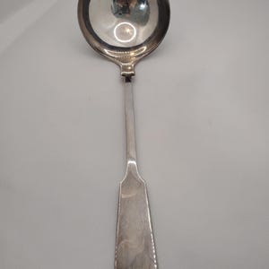 Könnte beinhalten: Eine silberne Schöpfkelle mit einer runden Schüssel und einem langen, schlanken Griff. Die Schöpfkelle besteht aus einem glänzenden, reflektierenden Metall. Der Griff hat ein flaches, längliches Ende. Die Schöpfkelle ist wahrscheinlich aus Edelstahl oder Silber.