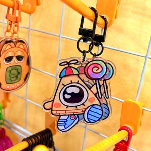 May include: A clear acrylic keychain featuring a triangular character with a single large eye, wearing a colorful propeller hat and blue sneakers, holding a multi-colored lollipop. The charm is attached to a black swivel clasp hook.