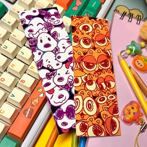 May include: Two colorful bookmarks with cartoon character designs. One bookmark features a pattern of white and purple cartoon faces, while the other has a pattern of orange cartoon faces. The bookmarks are next to a keyboard with custom keycaps.