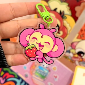 May include: A pink monkey-shaped keychain holding a red strawberry. The monkey has a yellow face and large pink ears. It is attached to a green clasp.