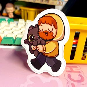 May include: A sticker featuring a cartoon character with a red beard and yellow hooded jacket, holding a black cat with large yellow eyes. The sticker is on a pink surface with a yellow basket in the background.