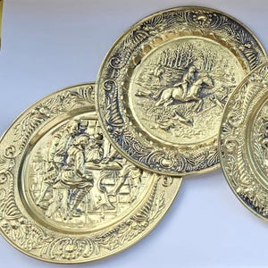 Könnte beinhalten: Satz von sechs goldfarbenen, dekorativen Metalltellern mit geprägten Designs. Jeder Teller zeigt eine andere Szene mit Figuren und Tieren.