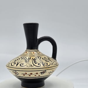 May include: A small black ceramic vase with a white and gold geometric design. The vase has a round body and a narrow neck with a handle.