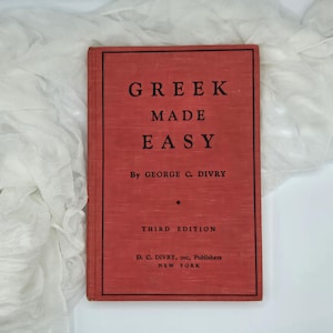 May include: A vintage book with a red cover, titled "GREEK MADE EASY". The author's name, George C. Divry, is printed in black. The book is a third edition, published by D. C. Divry, Inc., in New York. The book is a guide to learning Greek.
